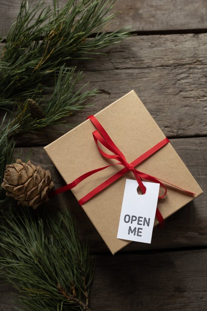A festive gift box with red ribbon and 'Open Me' tag on a rustic wooden background with pine sprigs.